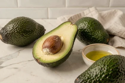 Avocat coupé en deux sur un comptoir en marbre blanc, révélant sa chair crémeuse vert pâle et son noyau brun, entouré d'avocats entiers et d'un bol d'huile d'olive